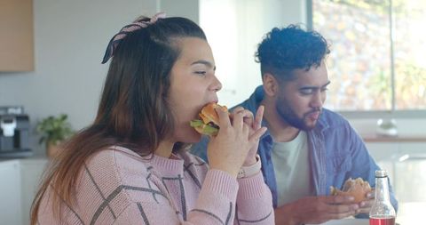 Diverse couple enjoying burgers at sunny kitchen counter, casual home meal moment