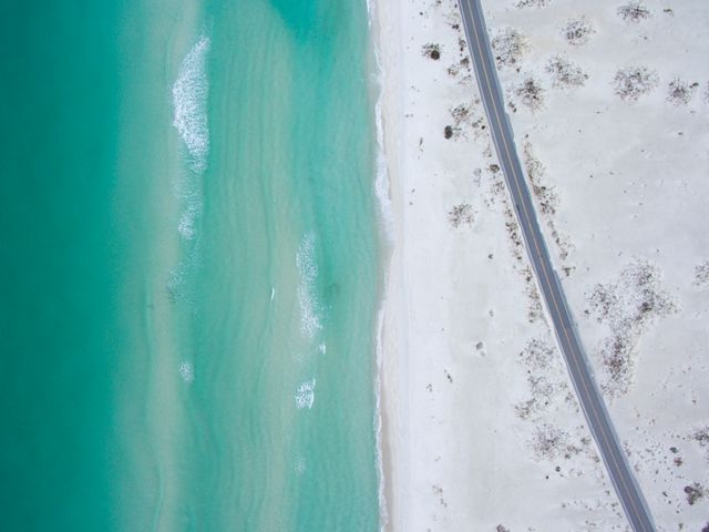Aerial photography depicting an idyllic beach with turquoise waters touching the sandy shore, alongside a serene coastal road. Beautifully showcases the tranquil environment, perfect for travel and tourism promotions, nature-inspired backgrounds, vacation brochures, and summer advertisements. It emphasizes the calm and natural beauty of coastal areas.