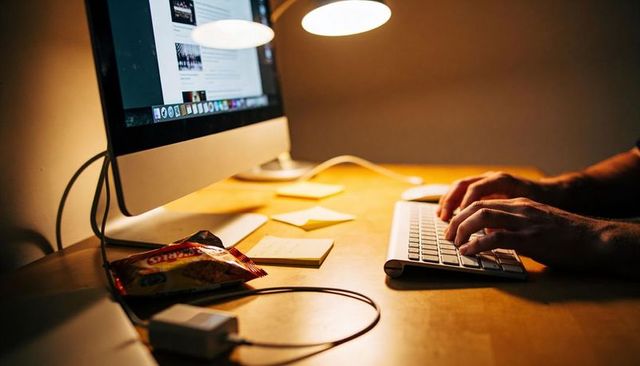 Late-night typing on wireless keyboard at minimal wooden desk with warm lamp glow
