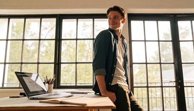Young designer leaning at studio workspace with laptop and sketchbook in sunlit loft
