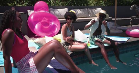 African american friends laughing on backyard pool edge with pink float and summer drinks