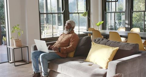 Senior Man Using Laptop Relaxing on Couch in Bright Living Room