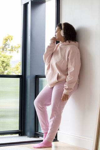 African american woman leaning by sunlit window listening with headphones in pink athleisure