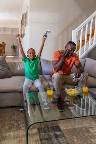 African american father and son celebrating gaming win on couch with snacks and juice