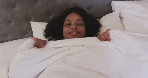 Smiling african american woman relaxing in bed under white duvet, cozy morning, minimalist bedroom