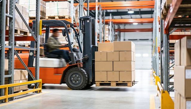 Forklift operator transporting pallet of cardboard boxes through busy warehouse aisle