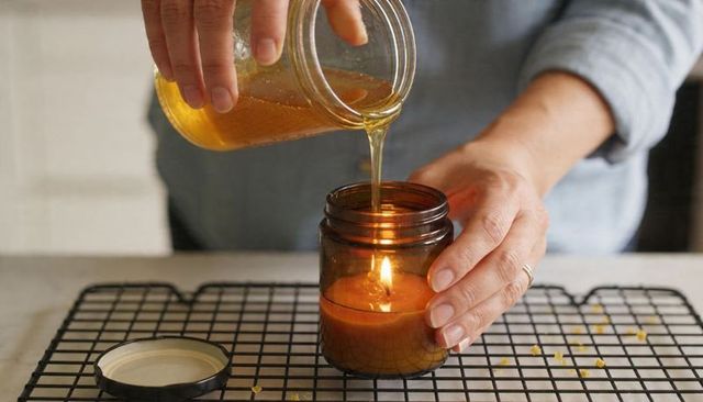 Pouring melted wax into amber glass candle jar with lit wick, hands holding jar