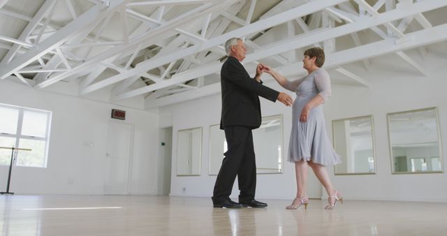 An elderly couple dressed in formal attire dances gracefully in a well-lit dance studio with large mirrors and a high ceiling. Perfect for scenes involving romance, fitness activities for seniors, or lifelong companionship. Useful in advertisements promoting active lifestyles, senior community activities, or dance classes.