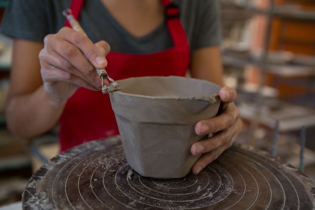 This image depicts a female potter molding a clay mug using a hand tool in a workshop. Ideal for use in articles or advertisements related to pottery classes, artisan crafts, creative hobbies, or handmade products. It can also be used in blogs or websites focusing on DIY projects, artistic endeavors, or showcasing the craftsmanship involved in pottery making.