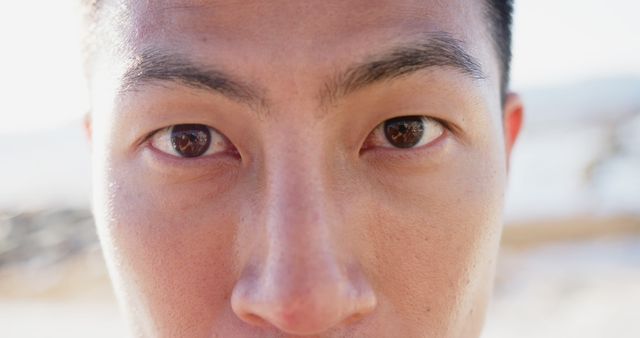 Close-up of an Asian man's face focusing intensely on the camera. His eyes are wide open and concentrated, reflecting determination and strength. Ideal for use in articles or campaigns about human emotions, determination, strength, or identity. Can also be used in healthcare materials to showcase eye health or vision-related topics.