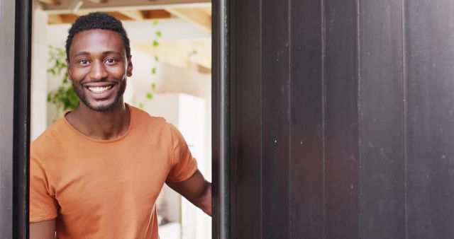 Man wearing orange shirt welcoming someone at the door with a warm smile. Can be used for concepts related to hospitality, welcome greetings, friendliness, personal interaction, and home lifestyle scenes.