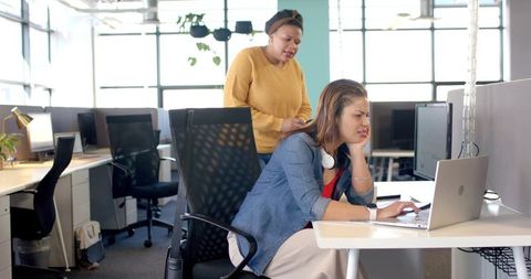 Female coworkers troubleshooting laptop in modern sunlit open-plan office