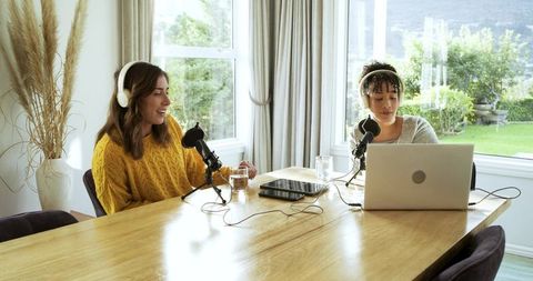 Two women recording podcast in sunlit home studio with laptop and desktop microphones