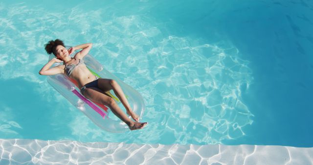 An overhead view of a woman lying on an inflatable raft in a clear blue swimming pool. Perfect for imagery related to summer, vacations, relaxation, leisure activities, tropical destinations, and sunny day scenes. Can be used in travel brochures, advertisements for resorts or pool-related products, and lifestyle blogs focused on summer activities.