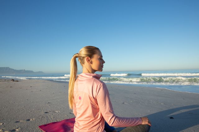 Caucasian woman wearing sports clothes, enjoying time at the beach on a sunny day, practicing yoga, sitting with legs crossed, meditating with eyes closed.