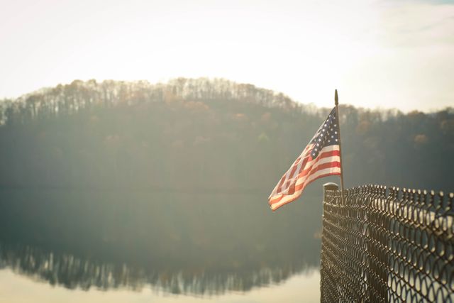 Displaying an American flag on a fence, this image portrays patriotism against a serene lakeside backdrop at sunrise. Ideal for themes related to national pride, tranquility, nature, morning scenes, and autumn landscapes. Useful for backgrounds, posters, website headers, and patriotic content.