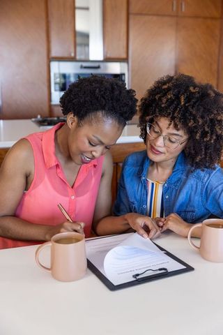 Signing and reviewing home paperwork together at kitchen island, female friends collaborating