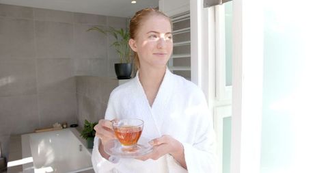 Woman in Bathrobe with Glass Teacup Enjoying Sunlit Bathroom