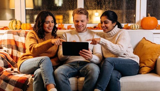 Friends sharing tablet on cozy autumn sofa with pumpkins, knit sweaters and warm blankets