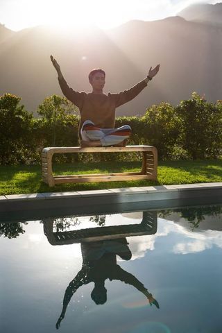 Young man meditating on wooden bench by pool at sunrise with mountain reflection