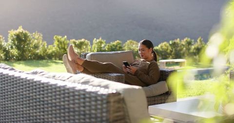 Woman relaxing on wicker sofa outdoors with smartphone