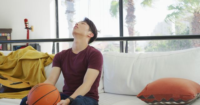 A young man sits on a white couch in a modern living room during the day, holding a basketball and looking up. Suitable for use in articles about stress, relaxation, or home lifestyle topics.