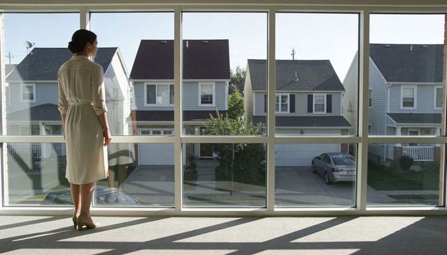 Woman standing at large window gazing at sunlit suburban street and houses