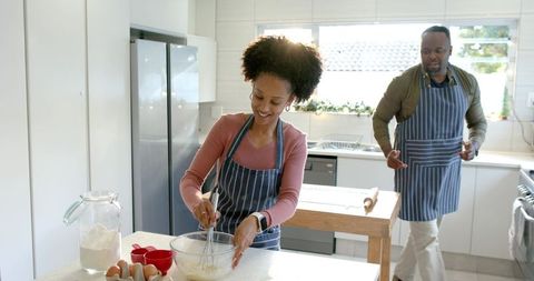 Diverse couple cooking together in bright modern kitchen mixing batter wearing striped aprons