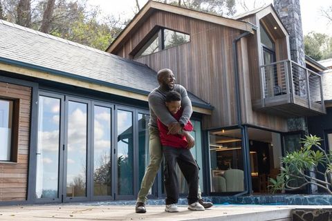 African american father hugging son on modern wooden deck laughing by glass doors