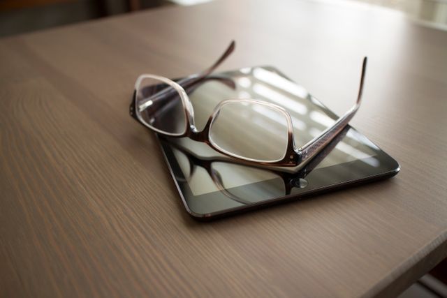 Close-up showing a pair of glasses lying on a tablet placed on a wooden desk. Useful for concepts involving technology, workspaces, digital reading, and study environments. Ideal for illustrating modern office or home office settings, emphasizing digital literacy and productivity.