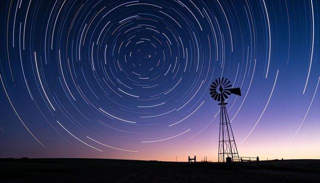 Silhouetted windmill under circular star trails at twilight over prairie
