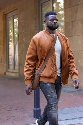 African american man walking in city wearing brown bomber jacket and crossbody bag