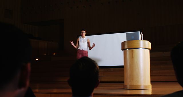 Female speaker is presenting on stage in an auditorium, using a projector and podium. This image is ideal for illustrating events such as business conferences, academic lectures, seminars, or professional training sessions. Could also depict public speaking, educational settings, and professional workshops.
