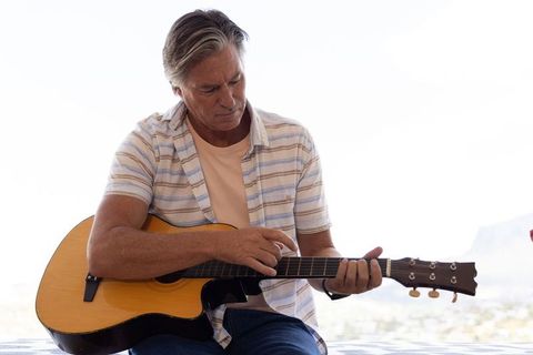 Mature man sitting on balcony plucking acoustic guitar, wearing wristwatch, sunny leisure