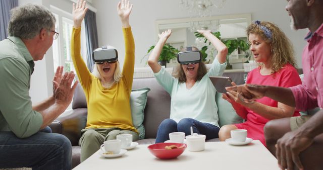 Seniors sitting on a sofa in living room, using virtual reality headsets, raising hands in excitement. Others are observing and using technology like tablets. The scene depicts an engaging and fun interaction with VR technology among elderly friends. Ideal for illustrating tech-savvy seniors, modern lifestyle, and social gatherings at home.