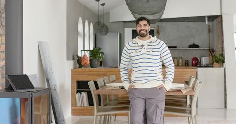 Smiling indian man standing in open-plan dining kitchen wearing striped sweater