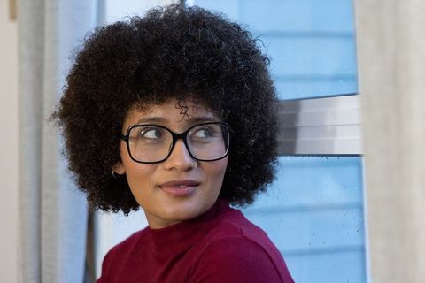 African american woman looking out rainy window wearing glasses and maroon sweater