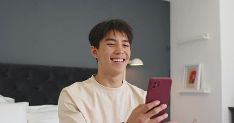 Smiling asian man holding red smartphone in modern bedroom using mobile for messaging