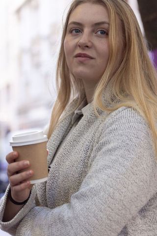Young woman holding coffee cup on city sidewalk wearing cozy gray coat, thoughtful look
