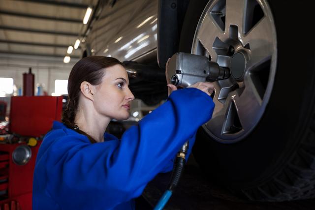 Female mechanic is fixing a car wheel with a pneumatic wrench at a repair garage. She is wearing a blue uniform and appears focused on her task. This image can be used to represent automotive services, gender diversity in technical professions, car maintenance tutorials, or promotional materials for repair shops and vocational training programs.