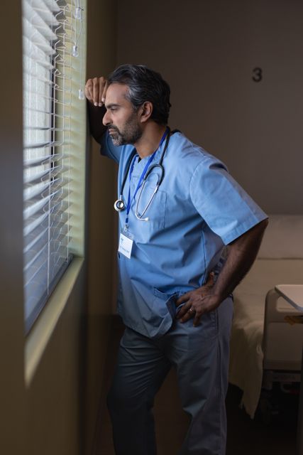 Biracial male surgeon in scrubs and stethoscope stands by window in hospital room, appearing thoughtful and contemplative. Useful for themes related to healthcare, medical professionals, hospital environment, and emotional moments in medical settings.