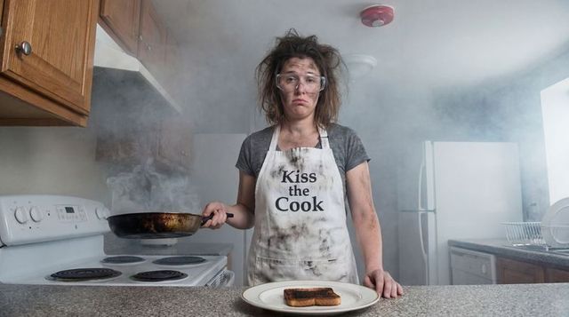 Stressed woman wearing kiss the cook apron and goggles holding burnt pan in smoky kitchen