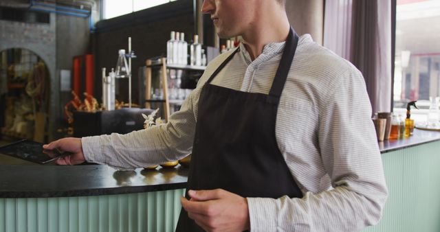 Barista wearing a black apron is taking an order using a digital tablet in a stylish modern cafe. The environment includes contemporary interior design features such as a sleek counter and various bottles in the background. This image can be used to depict themes related to customer service, technology in the hospitality industry, remote order systems, or modern coffee shop settings.