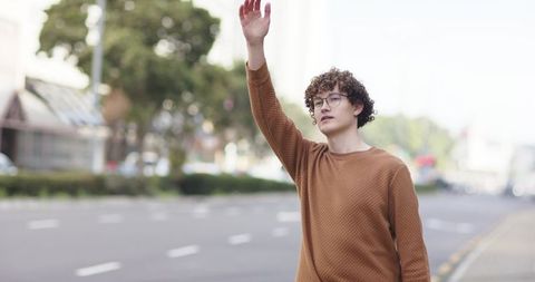 Curly-haired man hailing taxi on quiet city street wearing brown sweater and glasses