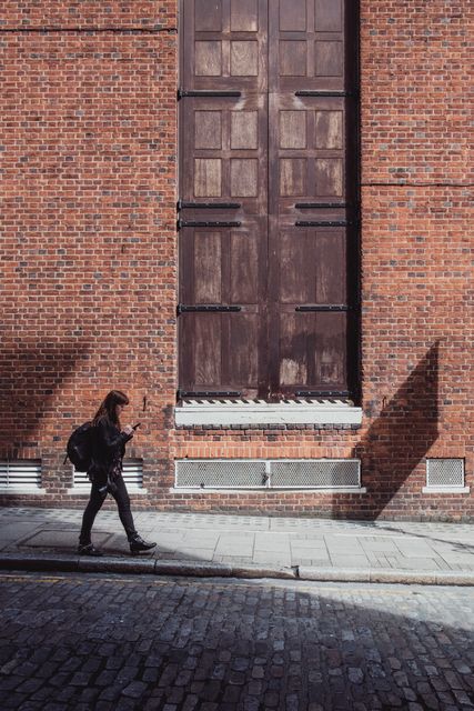 Young woman walking alone along a cobblestone street nearby a brick wall with large wooden doors. Ideal for themes related to urban exploration, everyday city life scenes, independence, and solitude in an urban environment. Useful for marketing materials, travel blogs, or advertisements promoting urban areas or pedestrian-centric imagery.