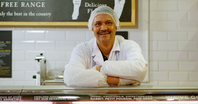 A smiling butcher dressed in a white coat and hairnet standing behind the counter with folded arms. Ideal for use in advertisements for butcher shops, meat markets, and food hygiene awareness campaigns.