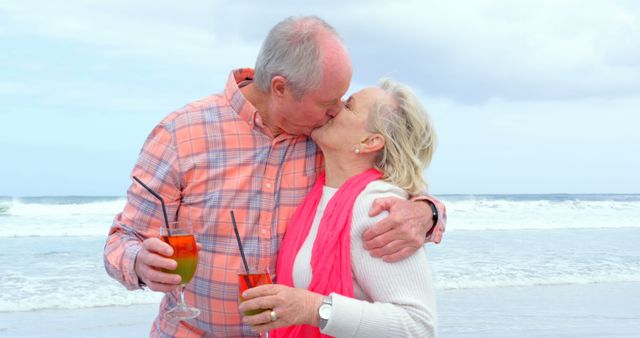 Happy senior caucasian couple kissing and holding cocktails on beach. Retirement, summer, leisure, vacation and senior lifestyle, unaltered.