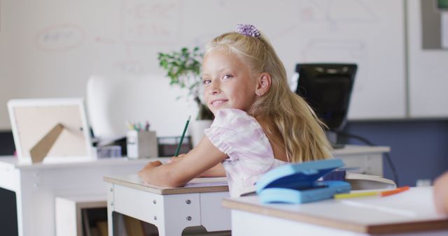 Young blonde girl smiling while sitting at her desk in a classroom environment. She is surrounded by typical educational tools and materials, suggesting a positive and engaging learning atmosphere. Can be used for educational websites, back-to-school promotions, or any context related to childhood education.