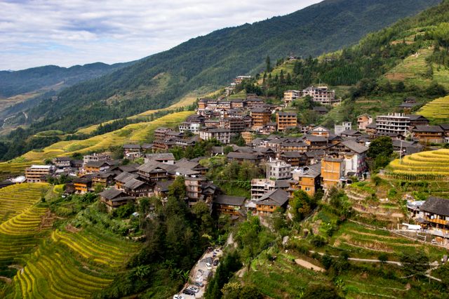 The image captures a traditional Chinese village nestled on a hillside, surrounded by verdant terraced rice fields. The picturesque rural landscape and unique architectural elements highlight the harmony between human settlement and nature. This visual is ideal for use in travel guides, cultural studies, agricultural features, and nature conservation campaigns.
