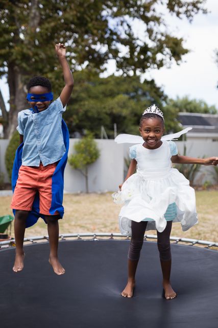 Two children dressed in costumes are jumping on a trampoline in a backyard. One child is dressed as a superhero with a blue cape and mask, while the other is dressed as a princess with a tiara and white dress. This image can be used for themes related to childhood, family activities, outdoor fun, imagination, and playful moments.